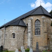 Kirche aus hellbraunen Steinen vor einer Grabesstätte unter blauem Himmel.