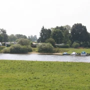 Campingplatz Fährhaus an der Weser in Thedinghausen Ein grüner Campingplatz an der Weser mit Wohnwagen, Booten und Bäumen unter bewölktem Himmel.