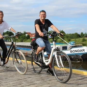 Zwei Menschen fahren auf Fahrrädern einen gepflasterten Weg entlang eines Flusses mit Boot im Hintergrund.