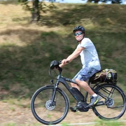 Radler Eine Person mit Helm und Sonnenbrille fährt bei Sonnenschein auf einem Elektrorad entlang einer grasbewachsenen Strecke.