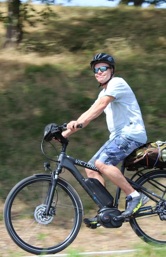 Radler Eine Person mit Helm und Sonnenbrille fährt bei Sonnenschein auf einem Elektrorad entlang einer grasbewachsenen Strecke.