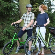 Radfahrer Zwei Menschen auf E-Bikes im Wald, lächelnd, bei sonnigem Wetter auf einem ruhigen Weg.