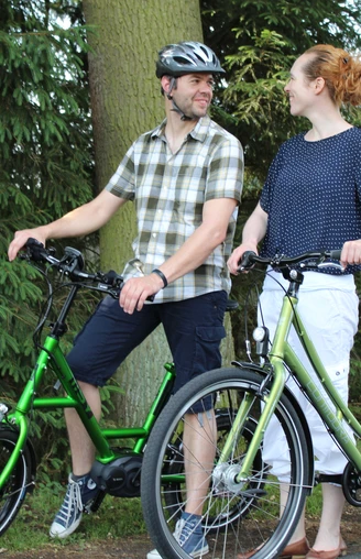 Radfahrer Zwei Menschen auf E-Bikes im Wald, lächelnd, bei sonnigem Wetter auf einem ruhigen Weg.