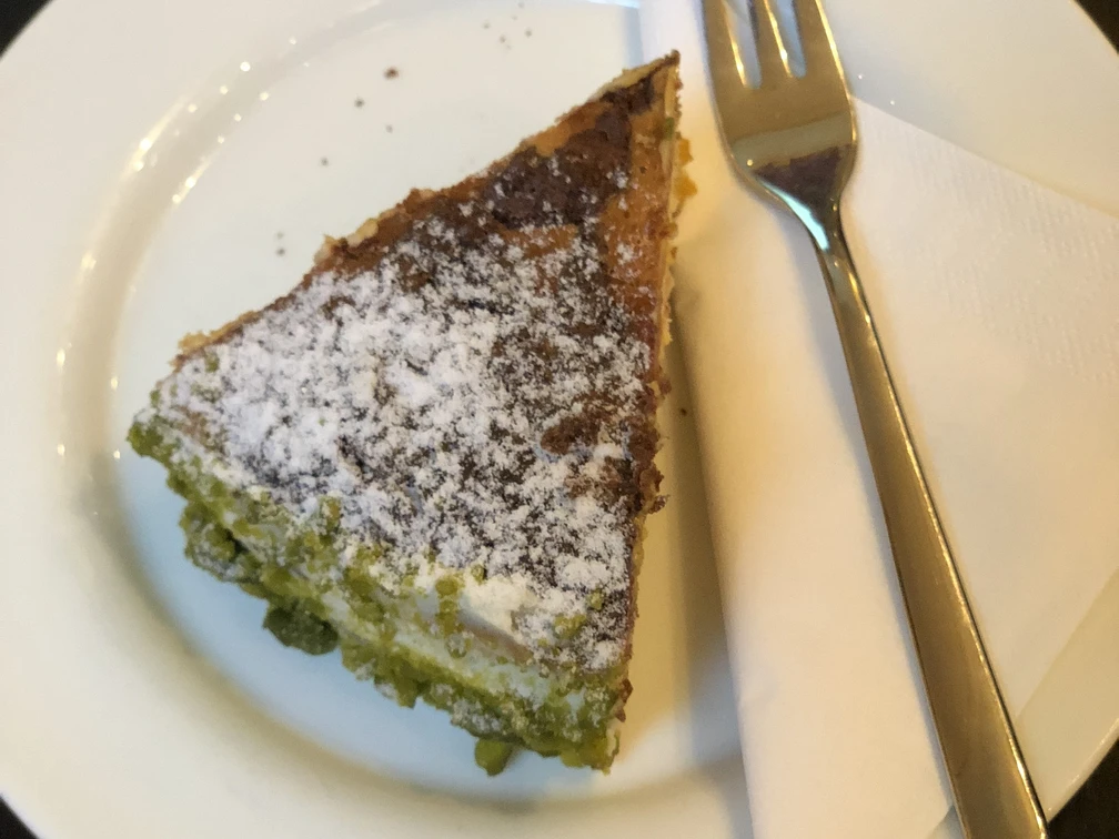 Ein Stück Torte mit grüner Füllung und Puderzucker, serviert auf einem weißen Teller mit Gabel.
