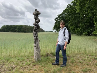 Löns Wanderweg Ein Mann steht glücklich neben einer kunstvoll geschwungenen Holzskulptur auf einem Feldweg.