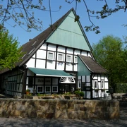 Altes Farmhaus Lienen_Frontansicht_Foto Dierkschneider.jpg Fachwerkhaus in Lienen mit grüner Markise, umgeben von Bäumen und einem gepflasterten Vorplatz.