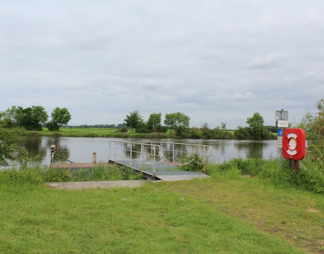 Landesbergen Schiffsanleger Ein Schiffsanleger an einem ruhigen Flussufer, umgeben von grüner Landschaft und bewölktem Himmel.