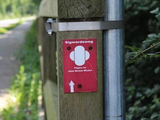 Markierung Pilgerweg Sigwardsweg Rotes Schild mit der Aufschrift "Sigwardsweg", befestigt an einem hölzernen Pfosten. Hintergrund unscharf.