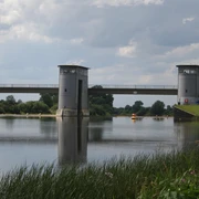 Intscheder Wehr Zwei Türme eines Wehrs reflektieren sich im stillen Wasser, flankiert von grünem Ufergras.