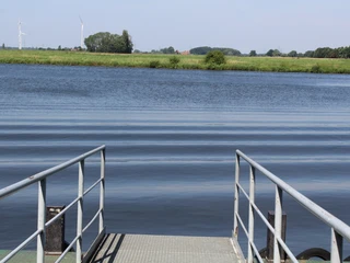 Anleger Dörverden Blick vom Anleger auf einen ruhigen Fluss, umgeben von grünen Wiesen und fernen Windrädern.