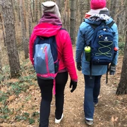 Zwei Personen wandern in warmer Kleidung über einen schmalen Waldpfad, umgeben von hohen Bäumen.