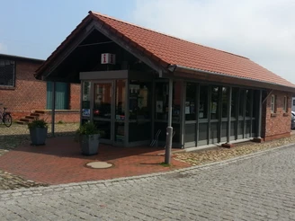 TourismusService Büro Einstöckiges Backsteingebäude mit roten Ziegeln, Glasfassade und Informationsschild an der Straße.