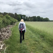 Wandern Eine Frau wandert auf einem grünen Pfad, umgeben von Feldern und Wäldern unter bewölktem Himmel.