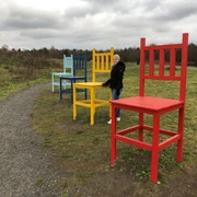 Kräher Höhe Bunte Riesenstühle auf einem Feldweg, Frau in Winterkleidung steht neben einem roten Stuhl.