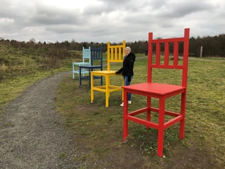 Kräher Höhe Bunte Riesenstühle auf einem Feldweg, Frau in Winterkleidung steht neben einem roten Stuhl.