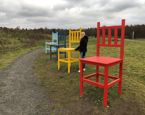 Kräher Höhe Bunte Riesenstühle auf einem Feldweg, Frau in Winterkleidung steht neben einem roten Stuhl.