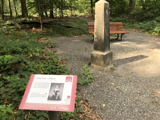 Hist. Promenaden Bad Rehburg Holzbank und Gedenkstein im Wald mit Infotafel über Marien-Platz auf einem Schotterweg bei Sonnenschein.