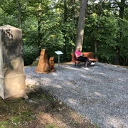 Eine Frau sitzt entspannt auf einer Bank in einem schattigen Wald, neben einer Holzskulptur.