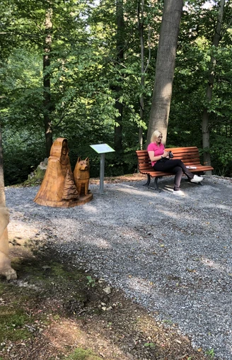 Promenaden Ernst August Höhe Eine Frau sitzt entspannt auf einer Bank in einem schattigen Wald, neben einer Holzskulptur.