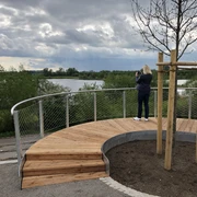 Eine Person genießt den weiten, wolkenverhangenen Blick über die Plattform auf einen ruhigen See.