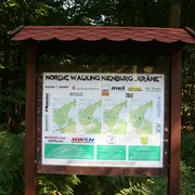 Nordic Walking-Schild im Wald mit Karte und Sponsorenlogos, umgeben von grünen Farnen und Bäumen.