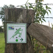 NW-Schild Baumpark Nordic-Walking-Schild an Holzzaun zeigt Richtung in grüner Landschaft mit Stacheldrahtumzäunung.