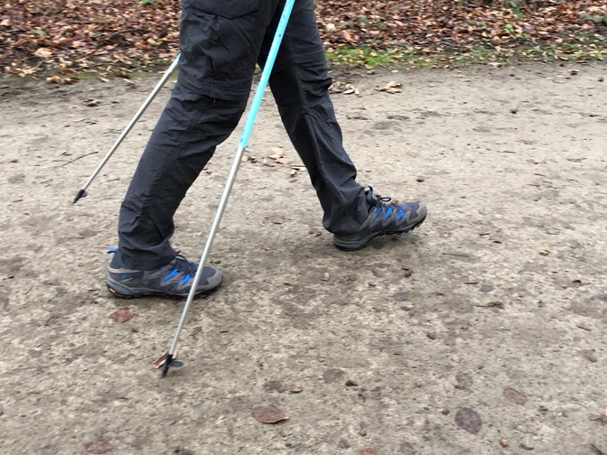 Nordic Walking Person auf Waldweg beim Nordic Walking mit Stöcken, herbstlicher Boden, Fokus auf Beine und Schuhe.