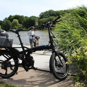 Radler Ein schwarzes Faltrad steht vor einem Flussufer, während ein Radfahrer im Hintergrund Wasser überquert.