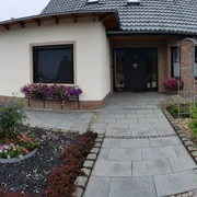 Gästehaus AM HERRMANN Zweistöckiges Gästehaus mit gepflegtem Garten, grauem Dach und Terrasse, umgeben von blühenden Beeten.