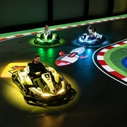 Ein Mann und zwei Kinder fahren in futuristischen Karts auf einer elektrischen Rennbahn. Bunte Lichter scheinen unter den Fahrzeugen auf, während sie über eine digitale Spielfläche manövrieren. Die Strecke ist mit farbenfrohen Markierungen und Symbolen gestaltet, was das Gefühl von Videospiel-Action in Echtzeit vermittelt. Spannung und Spaß sind in der elektrisierenden Atmosphäre spürbar, während die Fahrer durch die Kurven flitzen.