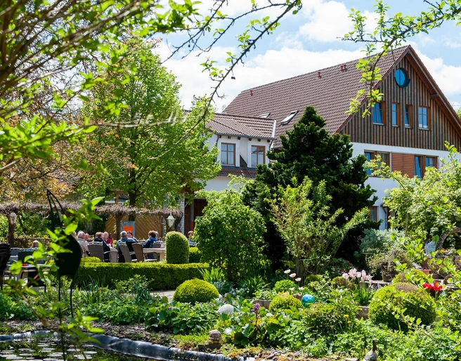 Landhaus Blumengarten: Außenansicht Üppige Gartenlandschaft mit blühenden Sträuchern umrahmt ein traditionelles Landhaus mit roten Ziegeldach.