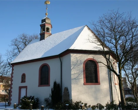 Schneebedeckte kleine Kapelle mit rotem Rahmen an Fenstern und Tür; umgeben von kahlen Bäumen.