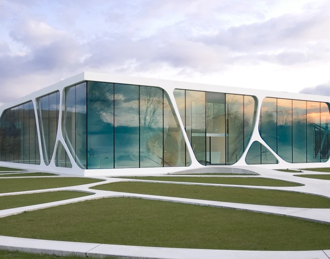 Modernes Gebäude mit großen Glasflächen und geschwungener Architektur in einer grünen Landschaft.