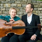 WEGE ERLEBEN Ein Mann und eine Frau sitzen vor einer Steinmauer, die Frau hält ein Cello in der Hand.