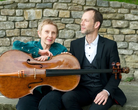 WEGE ERLEBEN Ein Mann und eine Frau sitzen vor einer Steinmauer, die Frau hält ein Cello in der Hand.