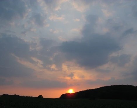 Sonnenuntergang Orange-roter Sonnenuntergang über bewaldetem Hügel, umgeben von dichten, grauen Wolkenformationen.