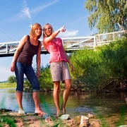 Erfrischung im Reich der Prachtlibellen Zwei Frauen barfuß in einem Fluss, eine zeigt lächelnd auf eine Brücke an einem sonnigen Tag.