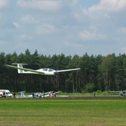 Segelflugplatz Segelflugzeug hebt vom grünen Flugplatz ab, im Hintergrund dichter Wald und ruhiger Himmel.