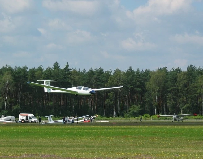 Segelflugplatz Segelflugzeug hebt vom grünen Flugplatz ab, im Hintergrund dichter Wald und ruhiger Himmel.