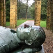 Ehrenmal Gedenkstätte mit liegender Steinskulptur, von Säulen umgeben, in einer bewaldeten Parklandschaft.