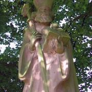 Das Nikolausdenkmal. Steinernes Nikolausdenkmal unter grünem Laub, zeigt Nikolaus mit Bischofsstab und Buch.