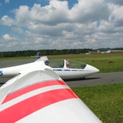 Segelflugplatz Ein Segelflugzeug am Boden, bereit zum Start, vor einer bewaldeten Landschaft und Wolken am Himmel.