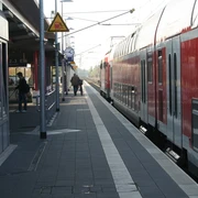 Bahnsteig Achim mit wartenden Passagieren und rotem Zug, eingefasst von Überdachung und Schienen.