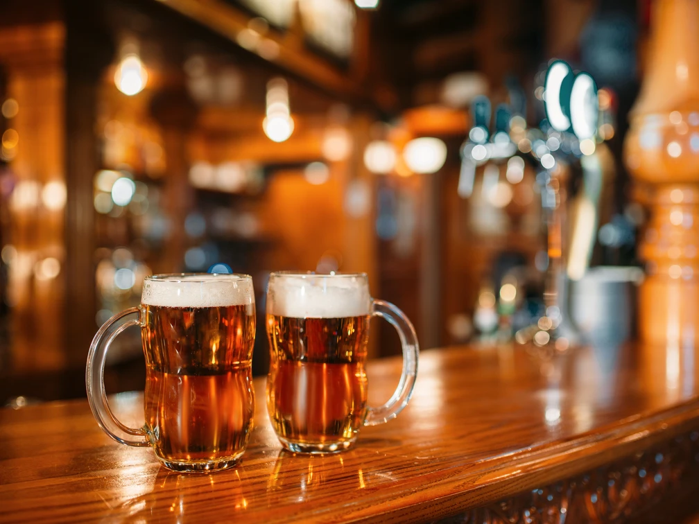 Two beer mugs on wooden bar counter, macro, nobody. Octoberfest symbol or concept. Glasses with golden beverage and foam on the table in pub, closeup view, blur background