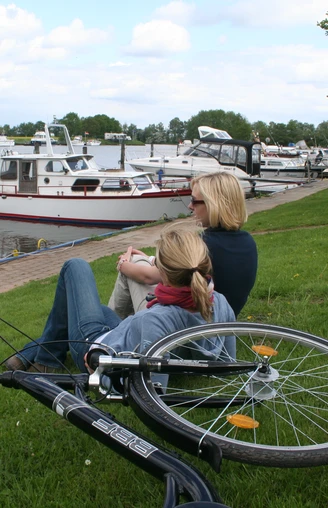 Zwei Frauen sitzen am Ufer eines Flusses mit Blick auf ankernde Boote, ein Fahrrad liegt daneben.