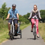 Zwei Radfahrer fahren auf einem asphaltierten ländlichen Weg in einer grünen, bewaldeten Landschaft.