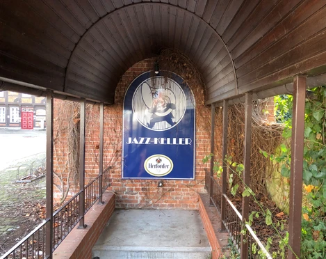 Eingang zum Jazz Keller Nienburg mit einem gewölbten Holzdach, umgeben von Backstein und grünen Pflanzen.