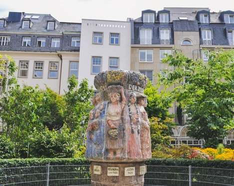 Women's fountain Der Frauenbrunnen zeigt zehn lebensgroße Frauenfiguren "die Frauen Kölns im Wandel der Zeiten“, umgeben von üppigem Grün, vor urbaner Kulisse. The Frauenbrunnen fountain shows ten life-size female figures "the women of Cologne through the ages", surrounded by lush greenery against an urban backdrop.