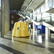 Das Puddingwunder in der Dr. Oetker Welt  Gelbe Puddingform-Skulptur in modernem Glasgebäude mit reflektierendem Boden und Treppen.