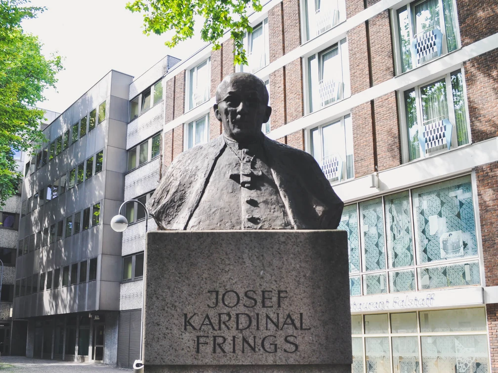 Joseph Frings Monument Das Joseph-Frings-Denkmal zeigt eine bronzene Büste des Kardinals auf einem steinernen Sockel, umgeben von modernen Gebäudeansichten.The Joseph Frings monument shows a bronze bust of the cardinal on a stone plinth, surrounded by modern views of buildings.